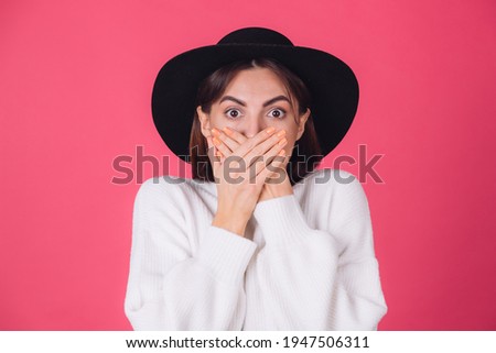 Stylish woman in white casual sweater and hat on red pink background copy space shocked look to camera cover mouth with hands