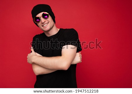 Photo shot of handsome joyful smiling young man wearing black t-shirt for mockup black hat and stylish sunglasses isolated over red background wall looking at camera