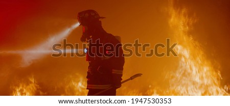 Dramatic silhouette of American firefighter in full gear exploring the huge fire zone Royalty-Free Stock Photo #1947530353