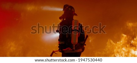 Dramatic silhouette of American firefighter in full gear exploring the huge fire zone Royalty-Free Stock Photo #1947530440