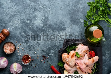 Raw chicken wings in metal pan or bowl with spices and ingredients for cooking on dark grey slate, stone or concrete background. Raw meat with spices at black table. Top view. Mock up.