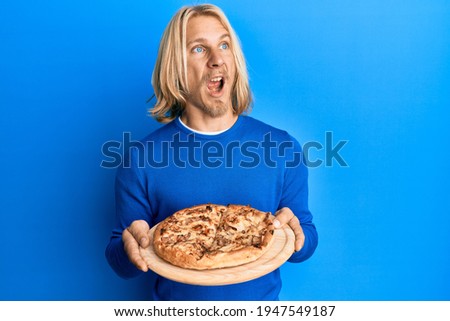 Caucasian young man with long hair holding italian pizza angry and mad screaming frustrated and furious, shouting with anger. rage and aggressive concept. 