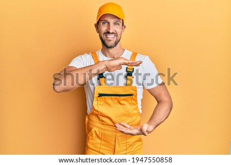 Young handsome man wearing handyman uniform over yellow background gesturing with hands showing big and large size sign, measure symbol. smiling looking at the camera. measuring concept. 
