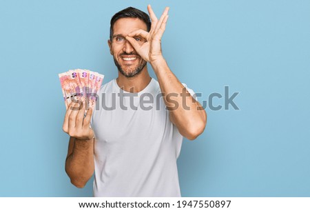 Handsome man with beard holding south african 50 rand banknotes smiling happy doing ok sign with hand on eye looking through fingers 