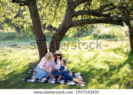 Multi-generation family spending time outdoors in sunny summer garden, sitting on checkered blanket under the big apple tree. Mature grandmother with daughter and granddaughter in park. Royalty-Free Stock Photo #1947563431