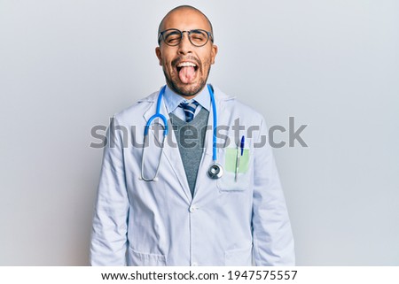 Hispanic adult man wearing doctor uniform and stethoscope sticking tongue out happy with funny expression. emotion concept. 