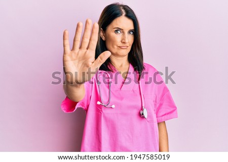 Middle age brunette woman wearing doctor uniform and stethoscope doing stop sing with palm of the hand. warning expression with negative and serious gesture on the face. 