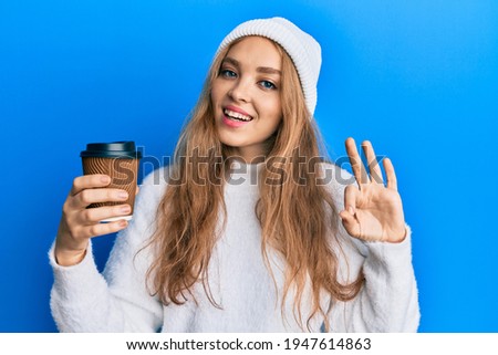 Beautiful blonde caucasian woman drinking a take away cup of coffee doing ok sign with fingers, smiling friendly gesturing excellent symbol 