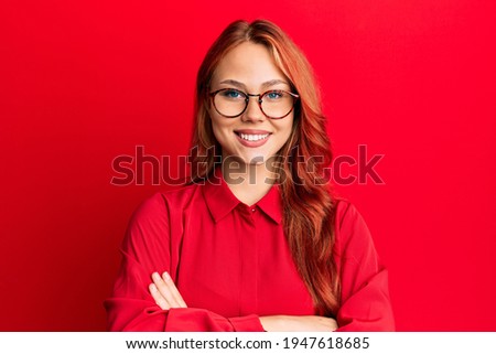 Young beautiful redhead woman wearing casual clothes and glasses over red background happy face smiling with crossed arms looking at the camera. positive person. 