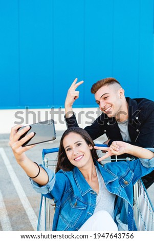 Lifestyle portrait of a young and beautiful couple having fun in a shopping cart and taking a selfie with a smartphone in blue background