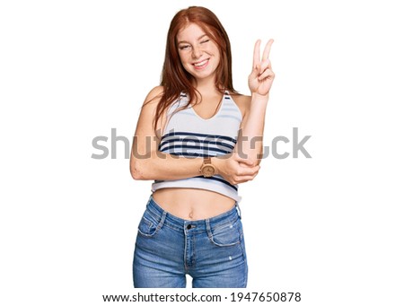 Young read head woman wearing casual clothes smiling with happy face winking at the camera doing victory sign. number two. 