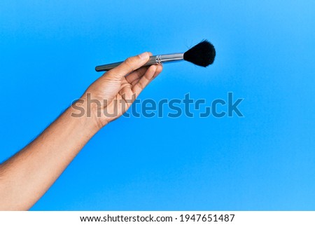 Hand of hispanic man holding makeup brush over isolated blue background.