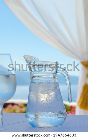 Close up of a jar and a glass of chilled water on a white table under a white curtain and an astonishing sea view on the front and a distant island.