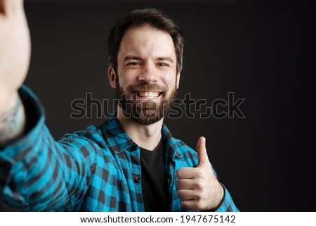 Bearded smiling man showing thumb up while taking selfie photo isolated over black background