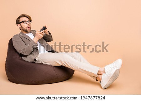 Full size photo of shocked amazed surprised man in glasses sit armchair playing playstation isolated on beige color background