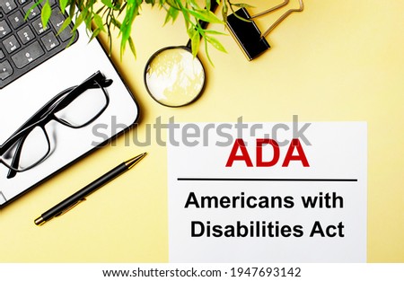 ADA Americans with Disabilities Act is written in red on a white piece of paper on a light yellow background next to a laptop, pen, magnifying glass, glasses and a green plant.