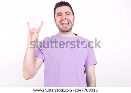 young handsome caucasian man wearing purple t-shirt against white background doing a rock gesture and smiling to the camera. Ready to go to her favorite band concert.