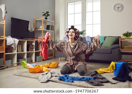Exhausted young woman cleaning her house. Desperate tired busy housewife in hair curlers and face mask picking up scattered clothes from floor, crying and complaining about terrible mess at home Royalty-Free Stock Photo #1947706267