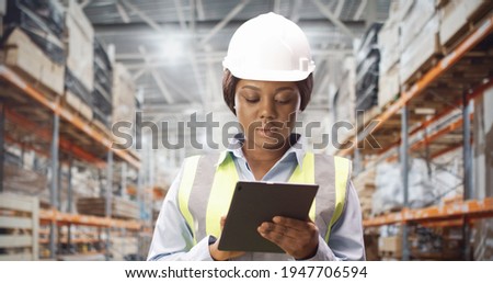African American female factory worker is wearing protection helmet typing on tablet device at warehouse factory. Woman industry worker browsing on device at work. Construction, engineer, close up