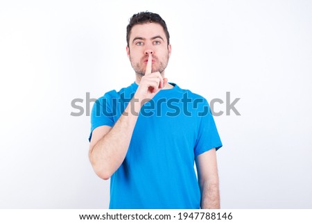 young handsome caucasian man wearing blue t-shirt against white background  makes silence gesture, keeps finger over lips. Silence and secret concept.