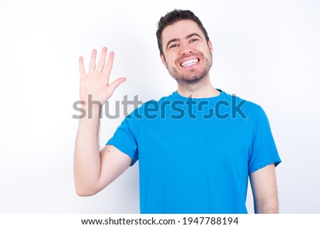 young handsome caucasian man wearing blue t-shirt against white background Waiving saying hello happy and smiling, friendly welcome gesture.