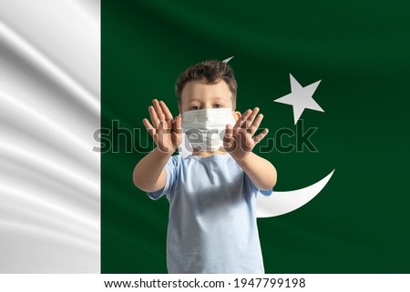 Little white boy in a protective mask on the background of the flag of Pakistan. Makes a stop sign with his hands, stay at home Pakistan.