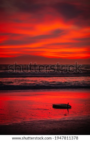 A wide shot of a bright red sunset with a surfboard on the beach in the foreground.
