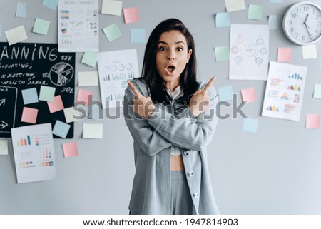 Beautiful, business girl on a gray work background shows index fingers up, arms crossed. Cute young woman in the office.
