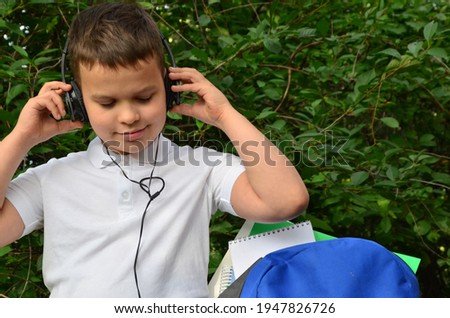 A schoolboy boy in a white shirt next to a briefcase in the park is playing music on headphones. Student learning listening audio tutorials in a park sitting on the grass in a park.