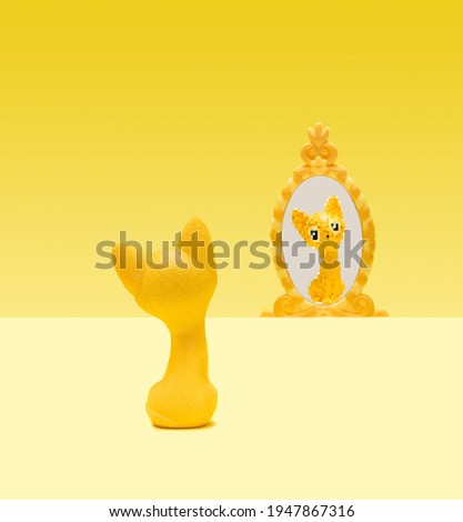 Single yellow cat toy seats on pastel yellow ground and looking itself in the mirror. In the reflection there is pixelated cat. Illuminate yellow background.