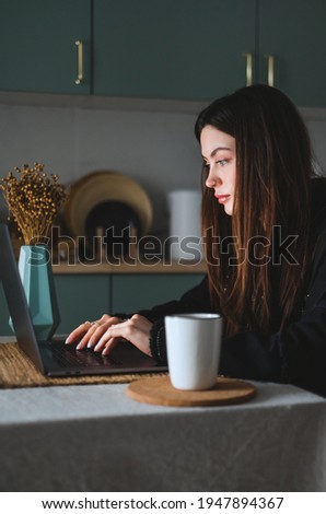 Young caucasian woman with brunette hair use laptop computer in the kitchen at home, typing text on the keyboard.