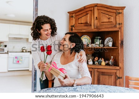 latin young woman and her mother middle age with flowers and gift box at home celebrating Happy mother's day in Mexico city