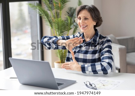 Happy deaf elderly woman uses sign language while video call using laptop while sitting in home office