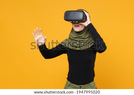 Amazed young arabian muslim woman in hijab black green clothes watch in vr headset gadget touch something like push click on button isolated on yellow background. People religious lifestyle concept