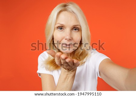 Close up of pretty elderly gray-haired blonde woman lady 40s 50s wearing white casual t-shirt doing selfie shot on mobile phone blowing sending air kiss isolated on orange background studio portrait