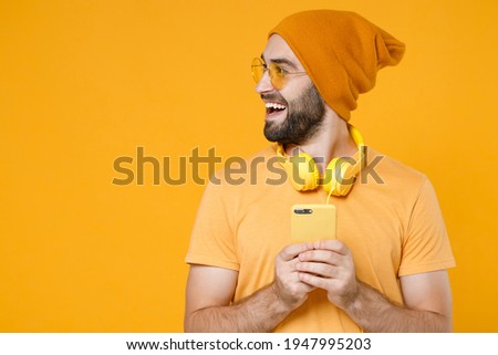 Cheerful young bearded man 20s wearing basic casual t-shirt headphones eyeglasses hat standing using mobile cell phone typing sms message looking aside isolated on yellow background, studio portrait
