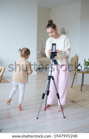 two sisters older and younger in pajamas at home shooting video on a phone with a tripod dressed in beige and white
