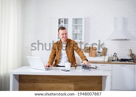 Handsome young businessman working with computer remotely sitting at the table in the kitchen. Nice happy man sitting in the room holding a drone in his hands