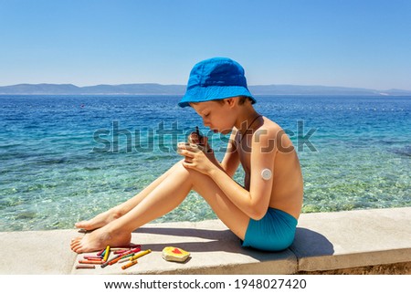Summer sea holidays: a boy with sugar monitoring system sitting on the beach and draws on pebbles. Concept life with diabetes, glycemic control. Children's creativity in the fresh air on vacation Royalty-Free Stock Photo #1948027420