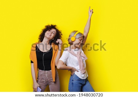 Young women are laughing and dancing on a yellow studio wall using headphones