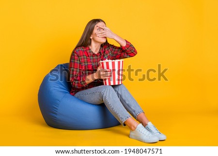 Full size photo of young scared afraid girl cover face sit armchair watch horror movie isolated on yellow color background