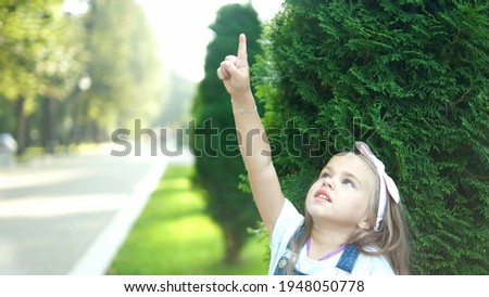 Pretty little child girl standing outdoors in green summer park pointing up with her finger.