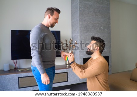 homosexual man kneeling down and asking his partner to marry him in the living room of his house. In the background you can see the gay pride flag.