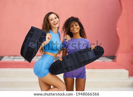 Two girls of different races on the background of a pink wall after training with sweat belts