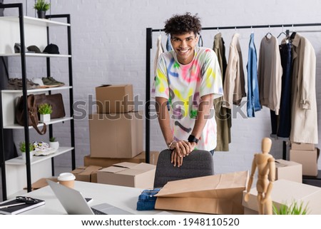 African american owner of showroom smiling at camera near shopping bag and box on table