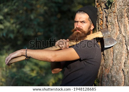 a serious brutal bearded man lumberjack with a long mustache in a warm hat holds an iron ax with two hands on a forest background