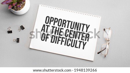 Opportunity at the center of difficulty is written in a white notebook next to a pencil, black-framed glasses and a green plant.
