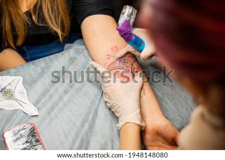Master tattoo draws the red paint on the clients tattoo. Tattoo artist holding a pink tattoo machine in black sterile gloves and working on the professional blue mat.