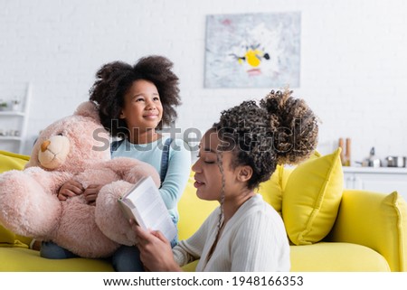 young african american woman reading book to child sitting on couch with teddy bear