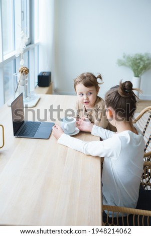 two sisters sit at home playing laptop games on the phone, watching cartoons on the laptop in a bright apartment in a sweater and light pajamas, drinking tea from a white cup